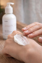 Leafia Skincare creamy white body lotion spread on back of hand, pump bottle softly blurred in background