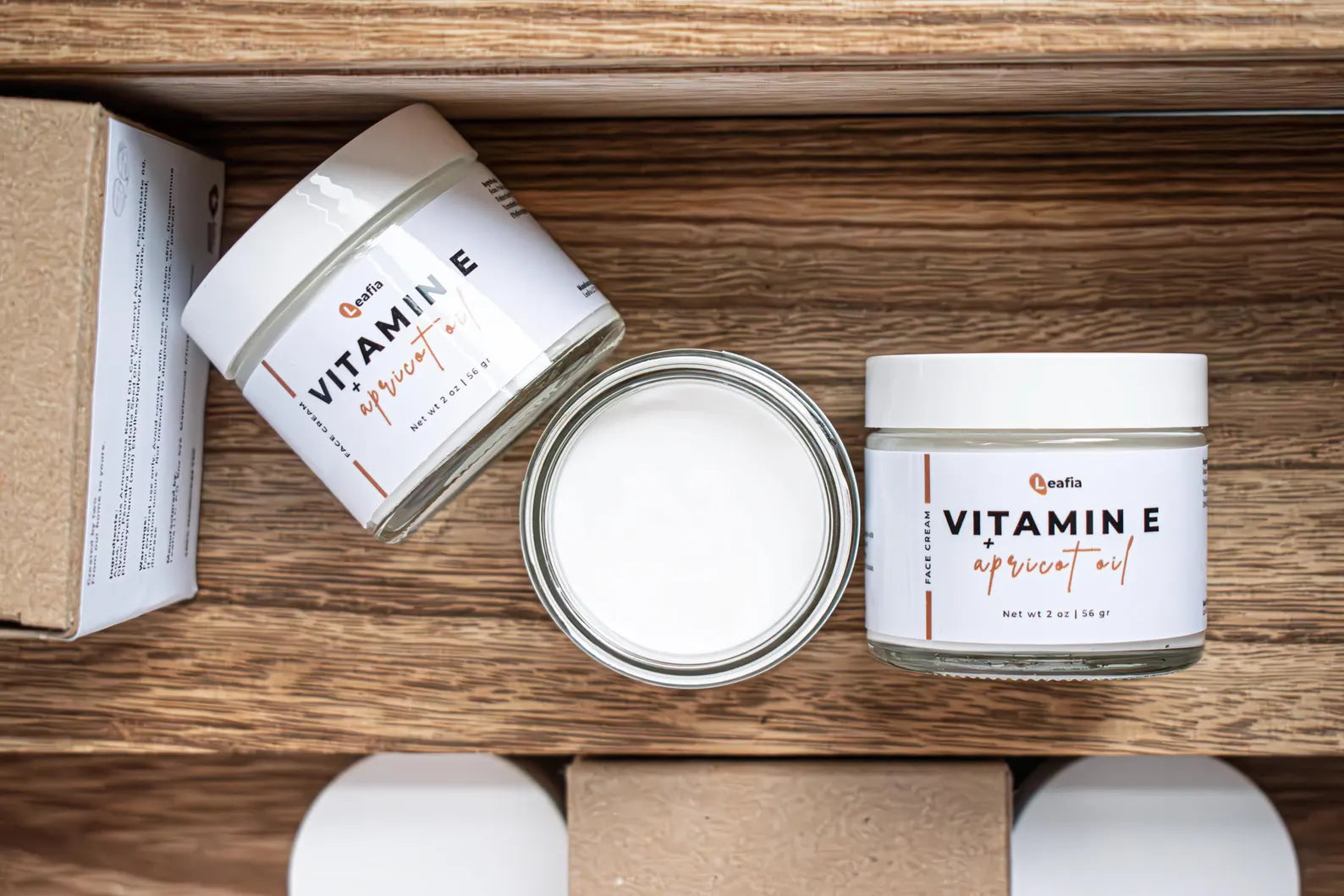 Leafia Skincare Vitamin E apricot oil lotion face and body moisturizer shown above with open jar, closed jar and packaging on natural wood