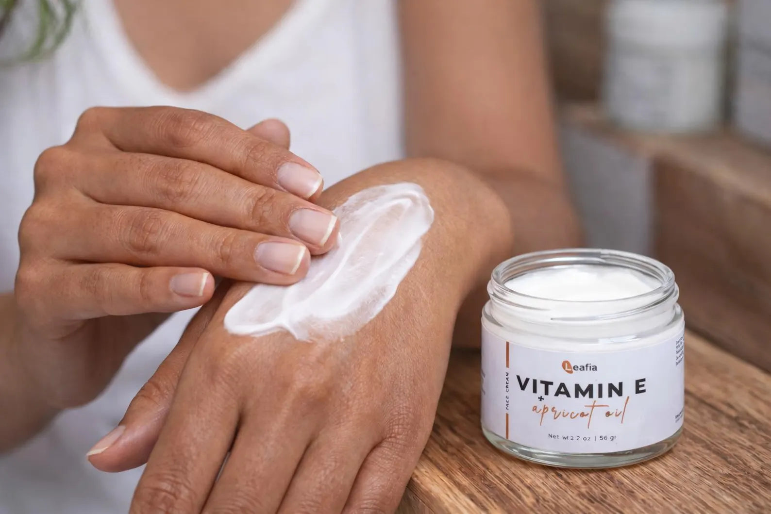 Hand applying Leafia Skincare Vitamin E apricot oil lotion, creamy lightweight texture from open glass jar on wood