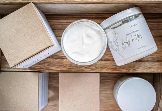 Leafia Skincare tallow body butter open from above, smooth creamy texture beside labeled jar and kraft box on wood