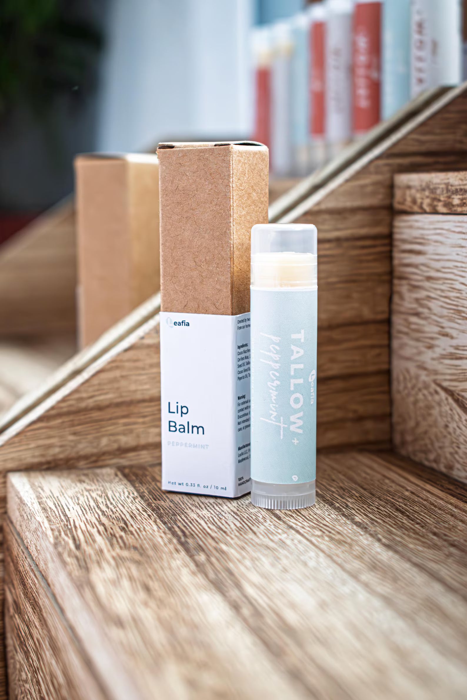 Leafia Skincare lip balm in natural twist-up tube beside kraft packaging on wood blocks with soft diffused light