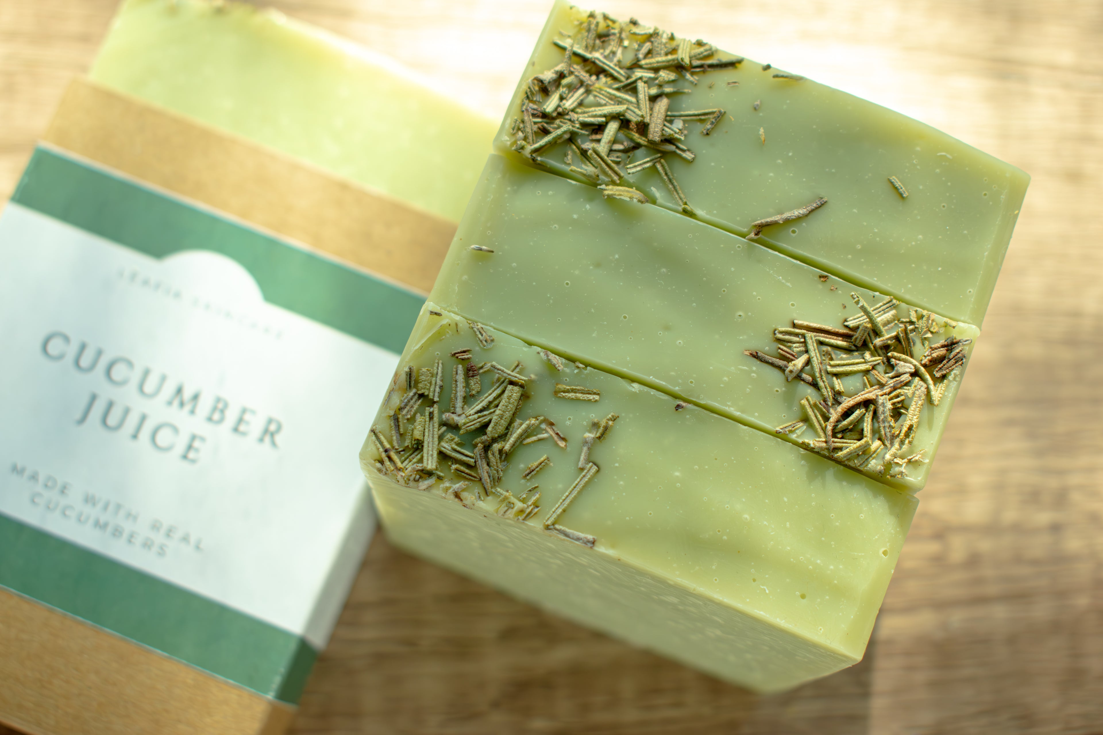 Green soap bar with visible herbs on a wooden surface, next to a label reading 'Cucumber Juice'.