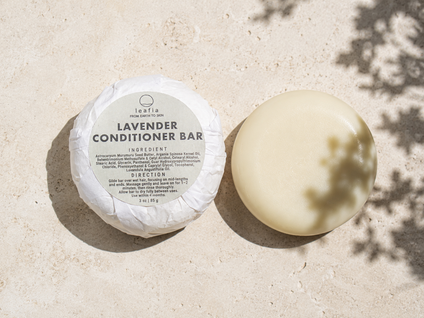 A round lavender conditioner bar shown wrapped in white paper with a printed label next to the unwrapped, solid cream-colored bar, placed on a light beige textured surface with dappled shadows.