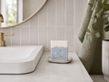 A rectangular bar of handmade soap with a white top layer and a blue marbled bottom layer sits on a small ceramic dish on a bathroom counter next to a sink.