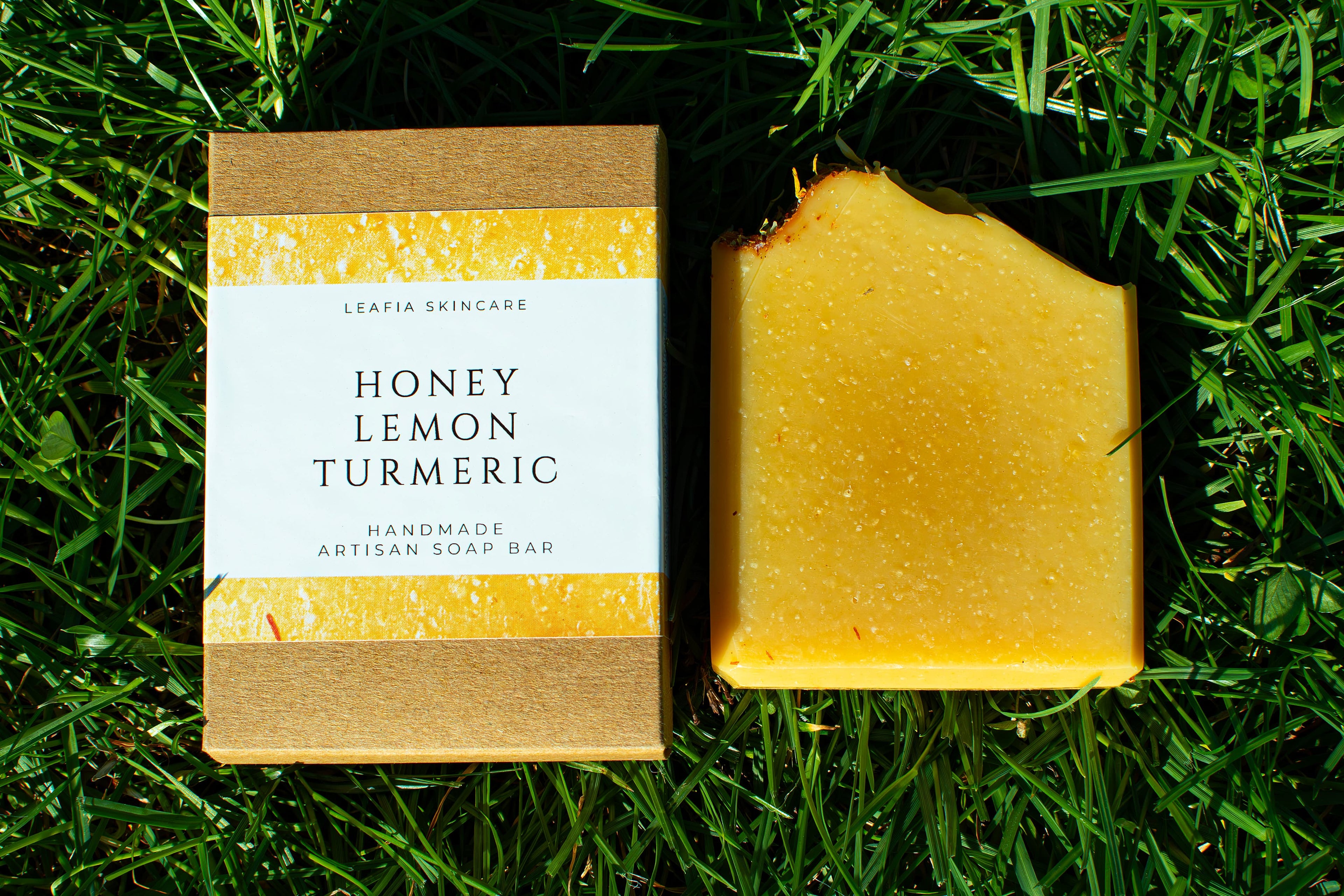 Orange soap on green grass. box label says honey, lemon, turmeric handmade artisan soap bar by leafia skincare
