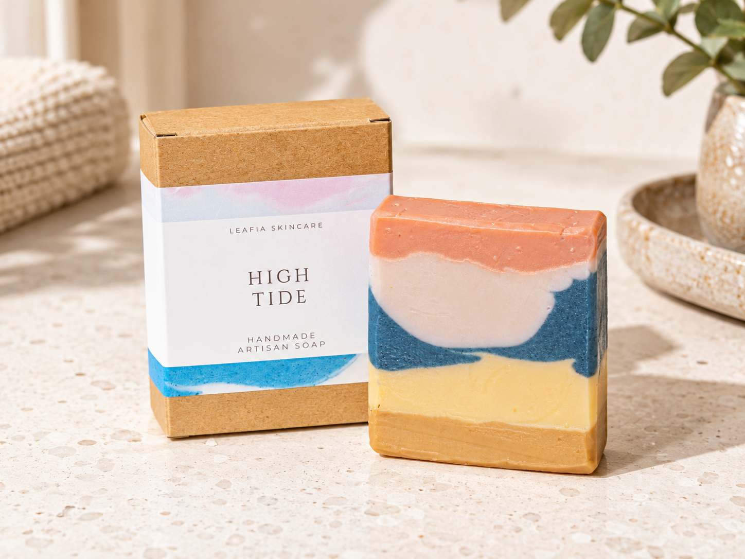 A handmade artisan soap bar with layered peach, cream, blue, and yellow stripes stands next to its kraft paper packaging box labeled 'HIGH TIDE' by Leafia Skinkare on a light stone surface.