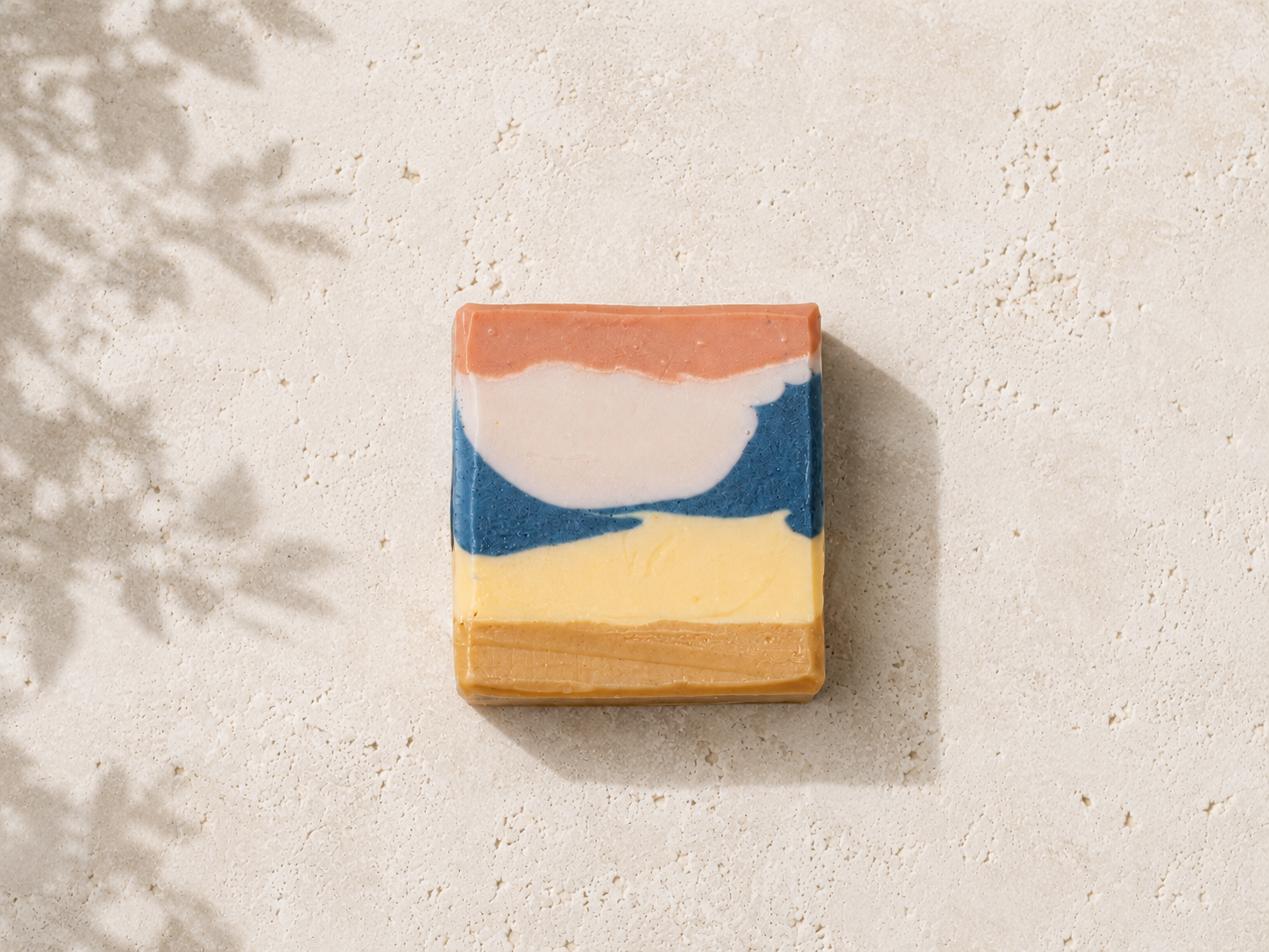A rectangular handmade soap bar with a layered abstract design in terracotta, cream, blue, and pale yellow, displayed on a textured light beige surface with soft leaf shadows.