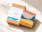 A handmade artisan soap bar with a marbled blue, white, peach, and yellow color pattern, displayed next to its kraft paper packaging box labeled 'HIGH TIDE' by Leafia Skincare on a light textured surface.