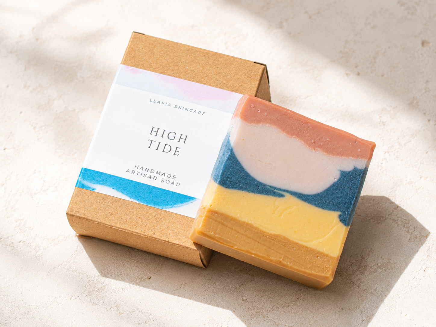 A handmade artisan soap bar with a marbled blue, white, peach, and yellow color pattern, displayed next to its kraft paper packaging box labeled 'HIGH TIDE' by Leafia Skincare on a light textured surface.