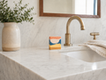 A close-up of a bathroom vanity featuring a white marble countertop and matching marble backsplash, with a brushed brass faucet, a folded cream towel, and a handmade art soap. A beige ceramic vase with green foliage sits on the left.