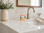 A close-up of a bathroom vanity featuring a white marble countertop and matching marble backsplash, with a brushed brass faucet, a folded cream towel, and a handmade art soap. A beige ceramic vase with green foliage sits on the left.