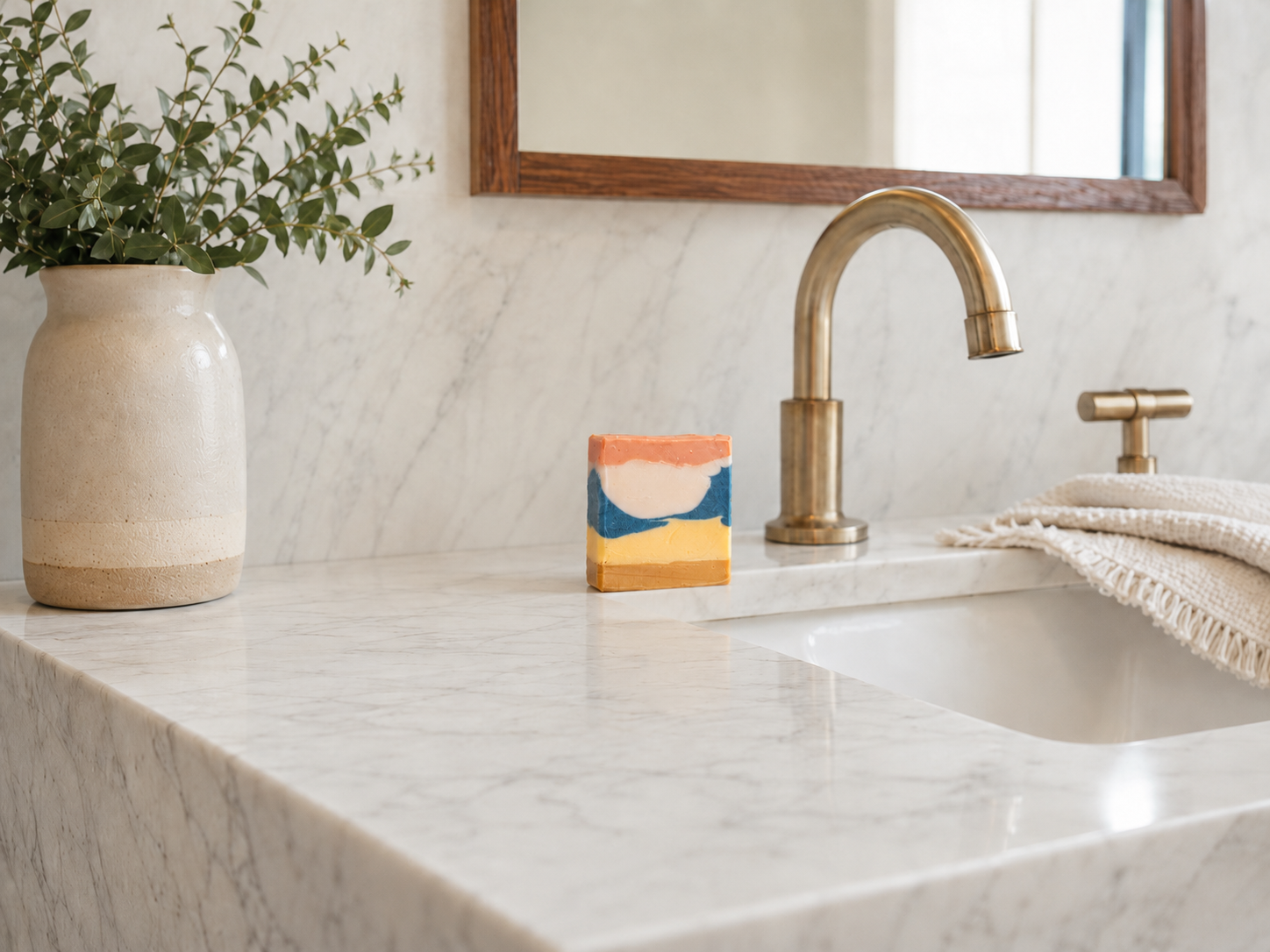 A close-up of a bathroom vanity featuring a white marble countertop and matching marble backsplash, with a brushed brass faucet, a folded cream towel, and a handmade art soap. A beige ceramic vase with green foliage sits on the left.