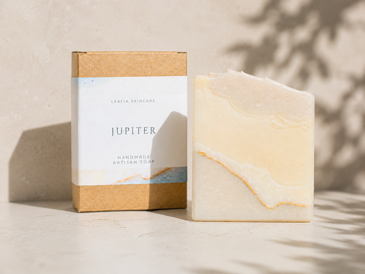 A bar of handmade artisan soap with a marbled cream and pale yellow pattern stands next to its kraft paper packaging box. The box features a white label with the brand name 'JUPITER' and 'LEAFIA SKINCARE' printed on it.