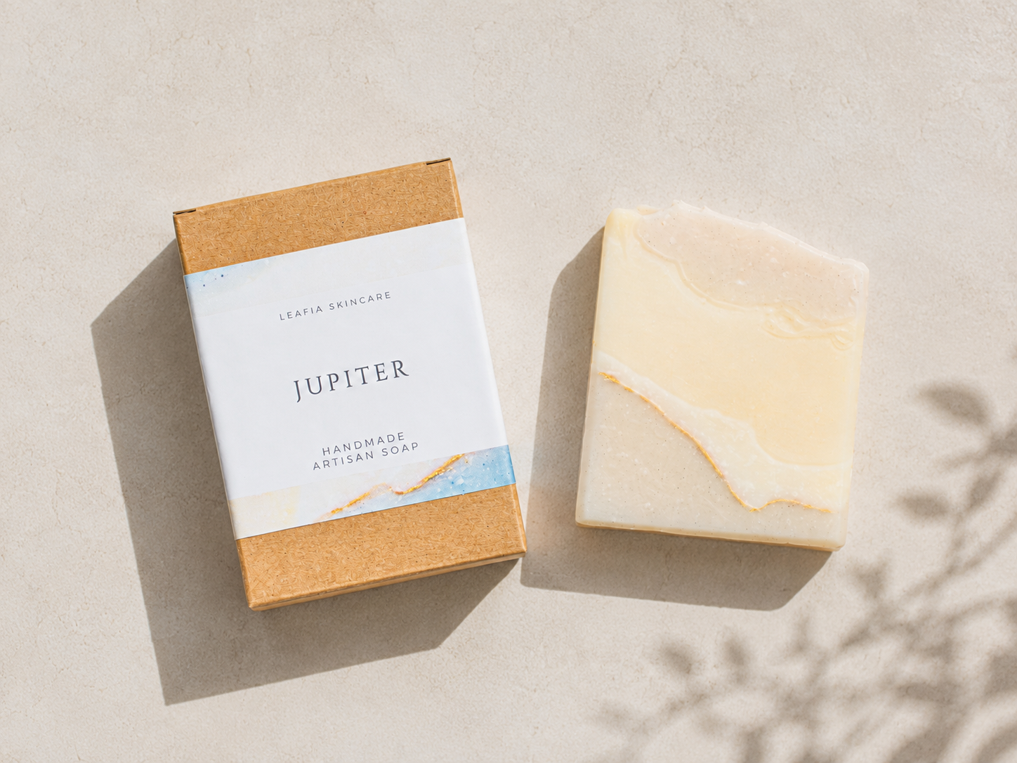A bar of handmade artisan soap next to its kraft paper packaging box, labeled 'JUPITER' by 'Leafia Skincare', on a light beige textured surface with soft leaf shadows.