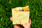 Hand holding a square piece of soap against a green grass background