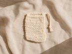 A small cream-colored crochet drawstring pouch with a tassel, laid flat on a draped beige linen fabric under soft natural light.