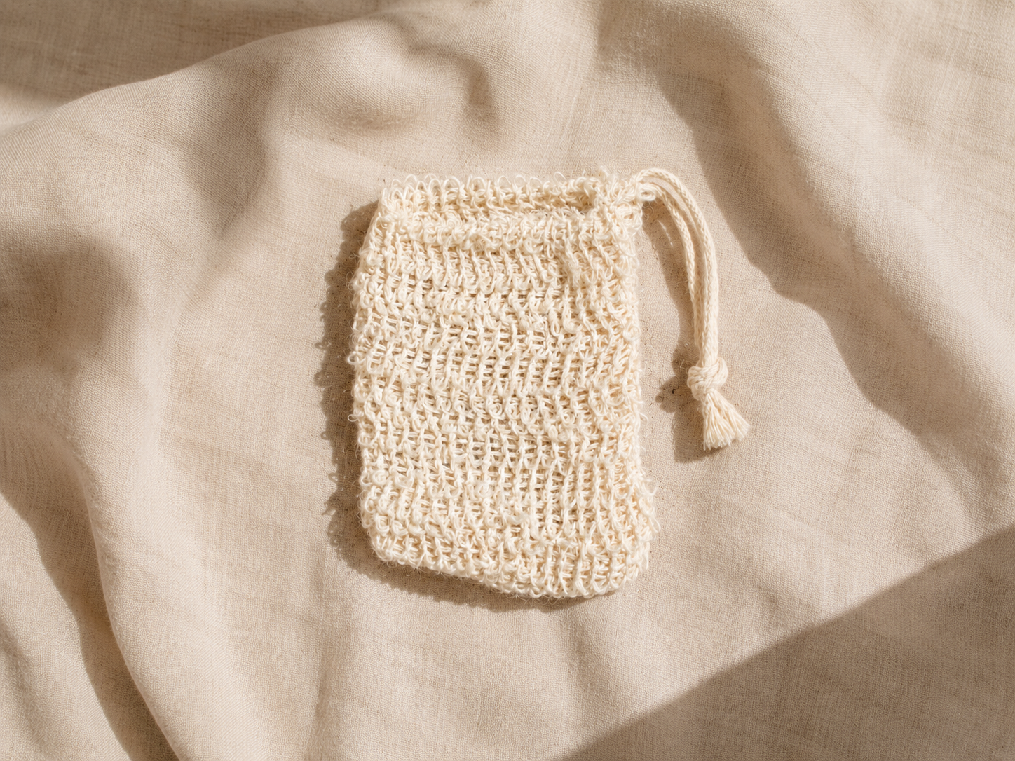 A small cream-colored crochet drawstring pouch with a tassel, laid flat on a draped beige linen fabric under soft natural light.