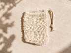 A small cream-colored crochet drawstring pouch with a tassel, laid flat on a textured beige surface with soft leaf shadows.