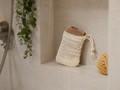 A cream-colored crochet drawstring bath bag holding a bar of soap, displayed on a beige tiled shower shelf next to a yellow sponge.