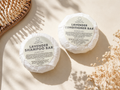 Two round wrapped hair care bars labeled Lavender Shampoo Bar and Lavender Conditioner Bar from the brand LEAFIA, arranged on a warm cream surface with dried flowers and soft shadows.
