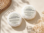 Two round wrapped hair care bars labeled Lavender Shampoo Bar and Lavender Conditioner Bar from the brand LEAFIA, arranged on a warm cream surface with dried flowers and soft shadows.