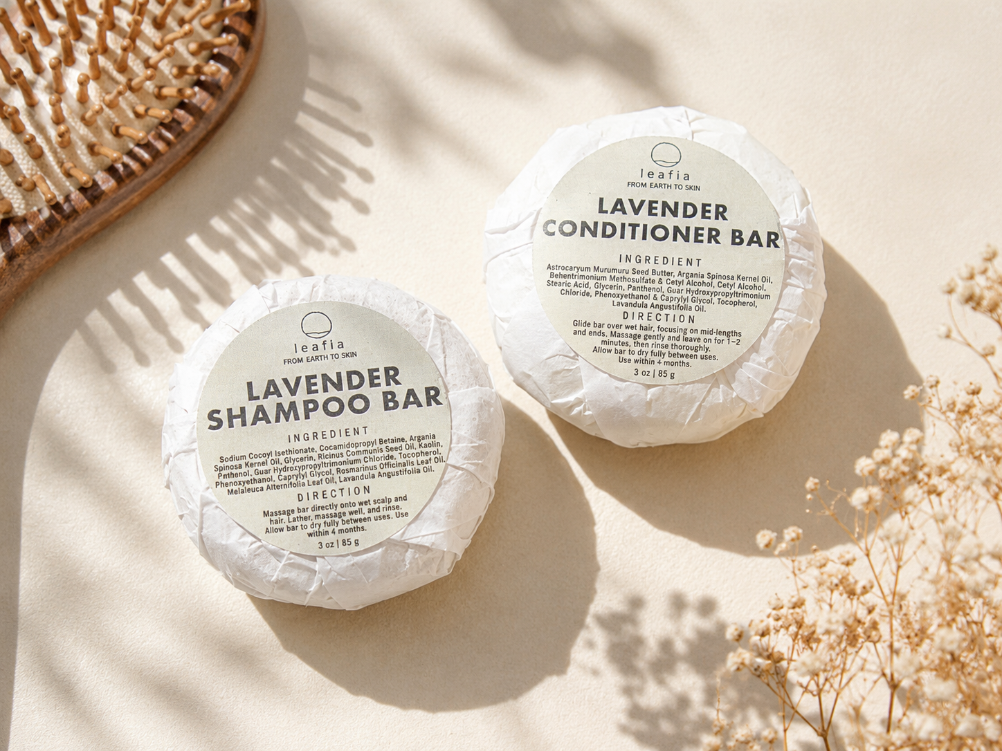Two round wrapped hair care bars labeled Lavender Shampoo Bar and Lavender Conditioner Bar from the brand LEAFIA, arranged on a warm cream surface with dried flowers and soft shadows.