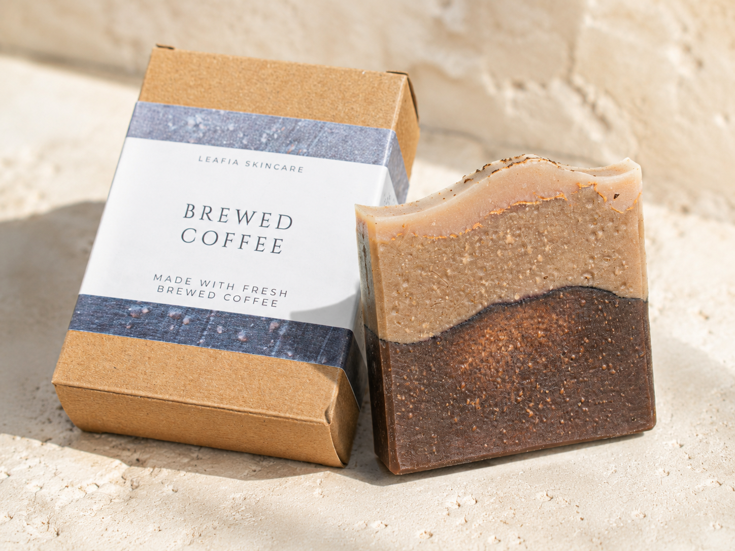 Bar of coffee-scented soap next to its packaging on a sandy surface