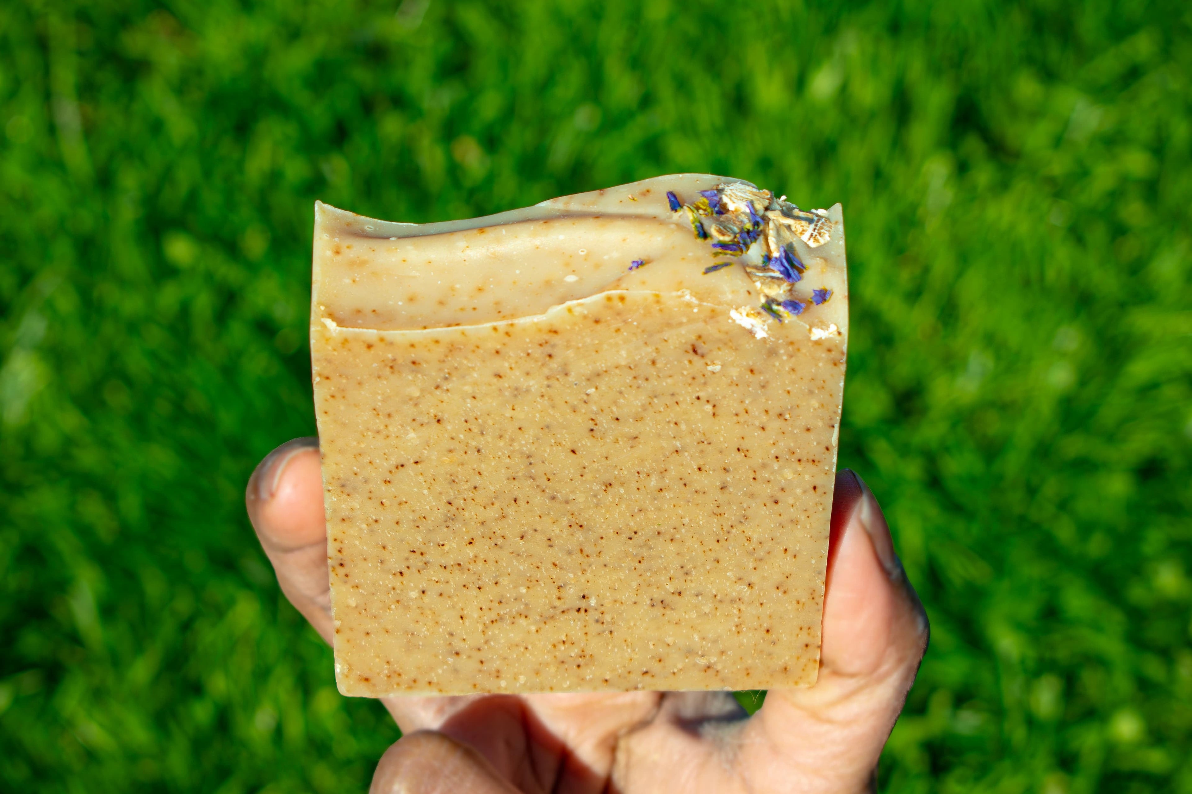 Hand holding a square piece of brown soap against a grass background