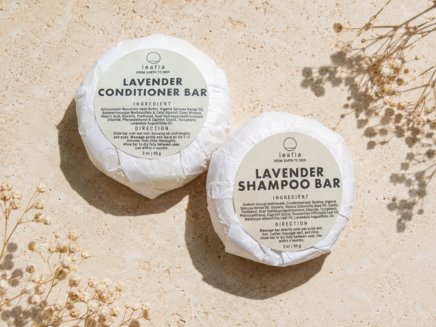 Two round wrapped soap bars labeled 'Lavender Conditioner Bar' and 'Lavender Shampoo Bar' from the brand ieafaa, arranged on a light beige textured surface with dried floral props and soft shadows.